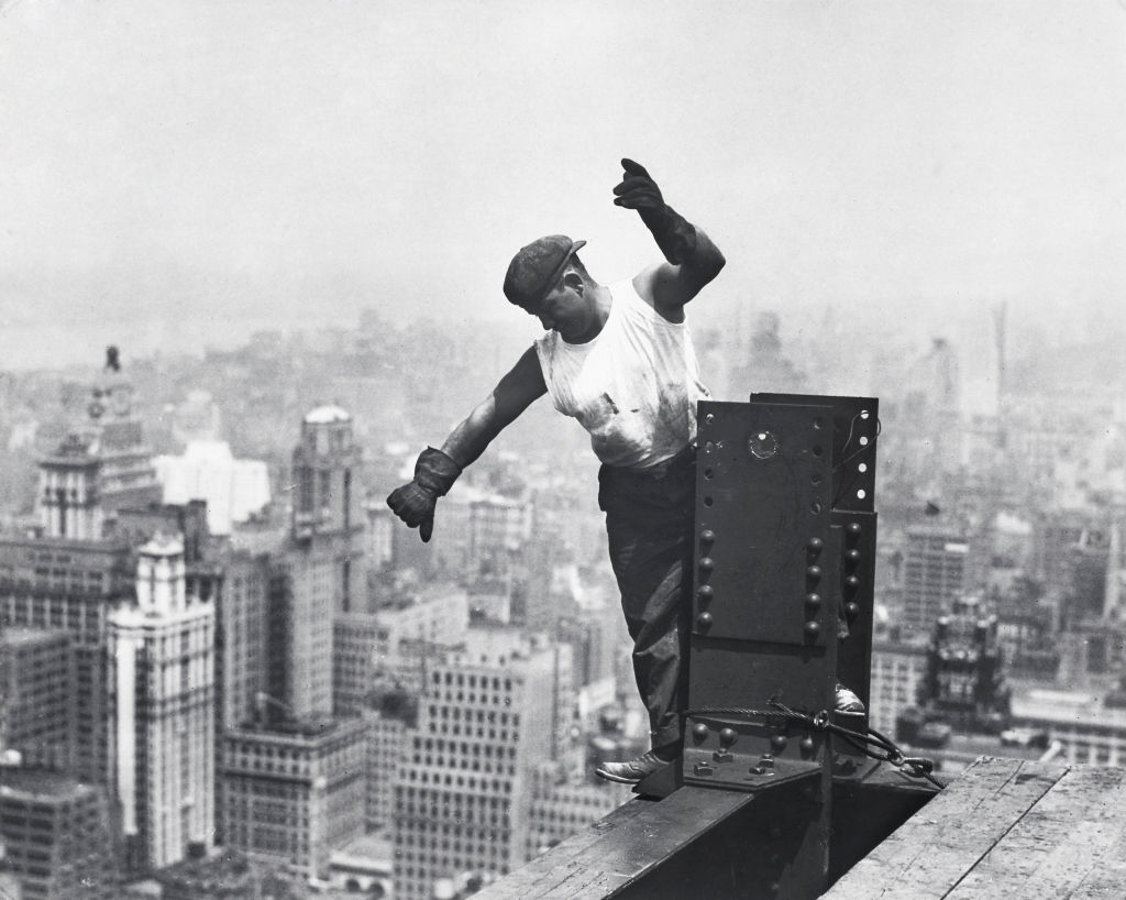 A construction worker balances on a steel beam high above a city, appearing to guide or signal something. He is wearing a flat cap, gloves, and a sleeveless shirt