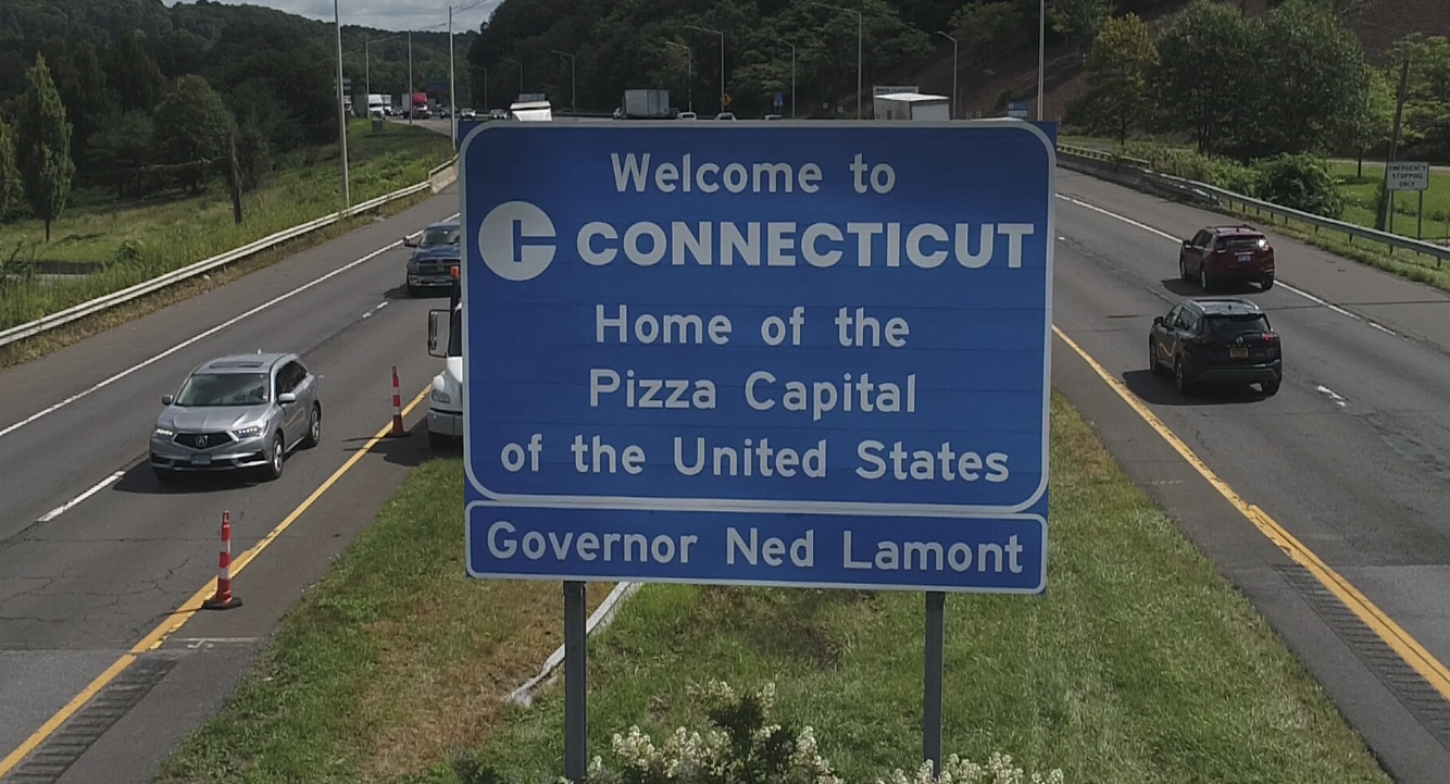 &quot;Welcome to Connecticut&quot; highway sign, reading &quot;Home of the Pizza Capital of the United States. Governor Ned Lamont&quot; with cars on the road in the background