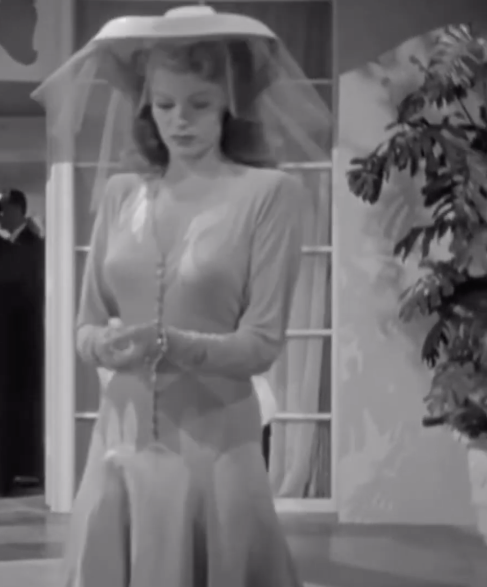 A woman dressed in a long-sleeved, button-down dress and a wide-brimmed hat with a veil, stands indoors, looking down at her hands
