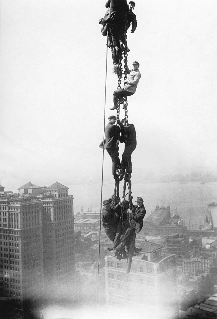 Historic photo of workers balancing on a tall steel beam, high above Manhattan during the 1930s construction boom. Rivers and buildings are seen below