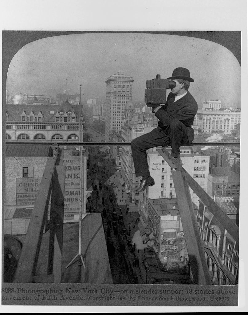 A man in a suit and bowler hat sits on a girder high above a city, using a large camera to photograph the New York City skyline. Text reads: "Photographing New York City—on a slender support 18 stories above pavement of Fifth Avenue."