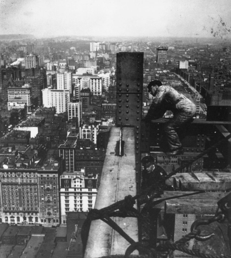 A construction worker in overalls stands on a steel beam high above a city, possibly during the construction of a skyscraper, with many buildings visible below