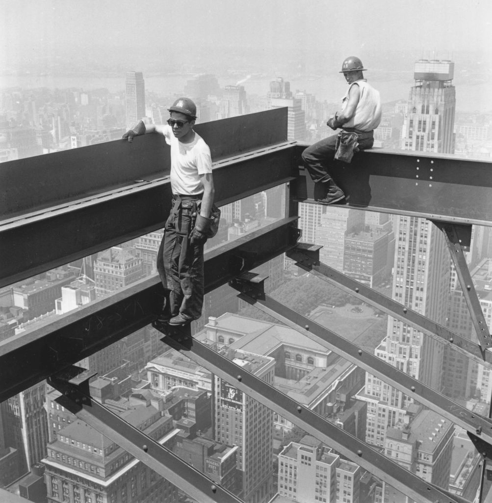 Two construction workers stand on steel beams high above a city, overseeing a building site