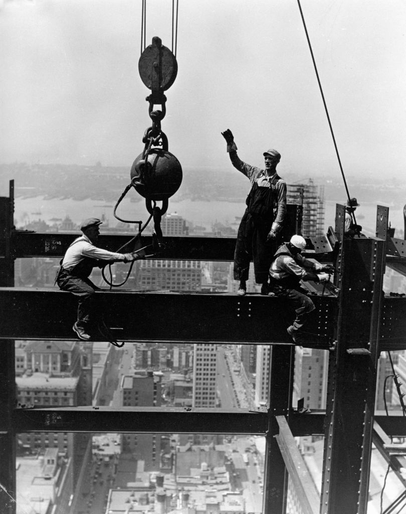 Construction workers on steel beams, high above a cityscape, guide a hoisted steel ball. The scene depicts early 20th-century high-rise construction