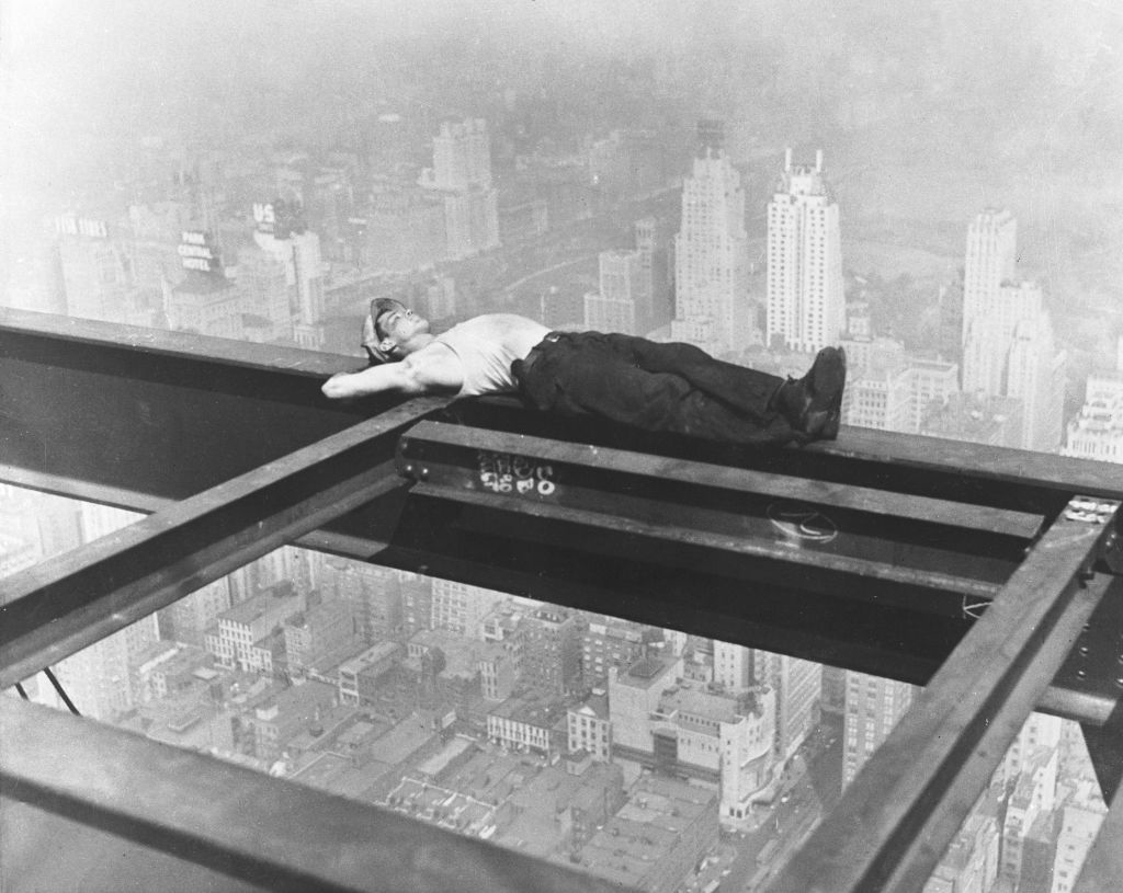 A construction worker is lying down on a steel beam high above a cityscape in a black-and-white photo, appearing to take a break