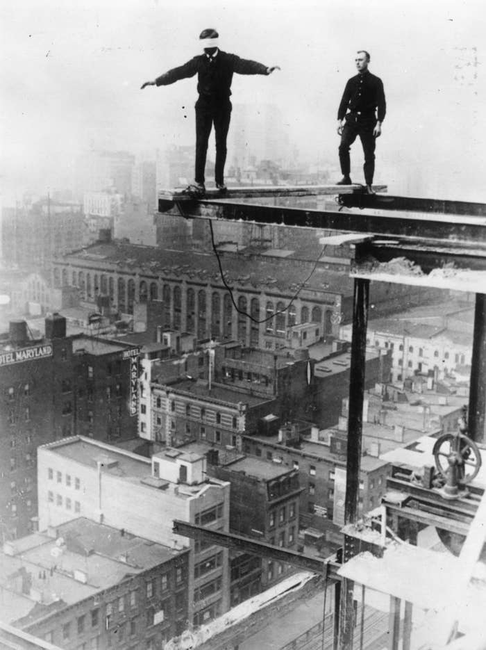Two men balancing on steel beams high above a city skyline during a construction project