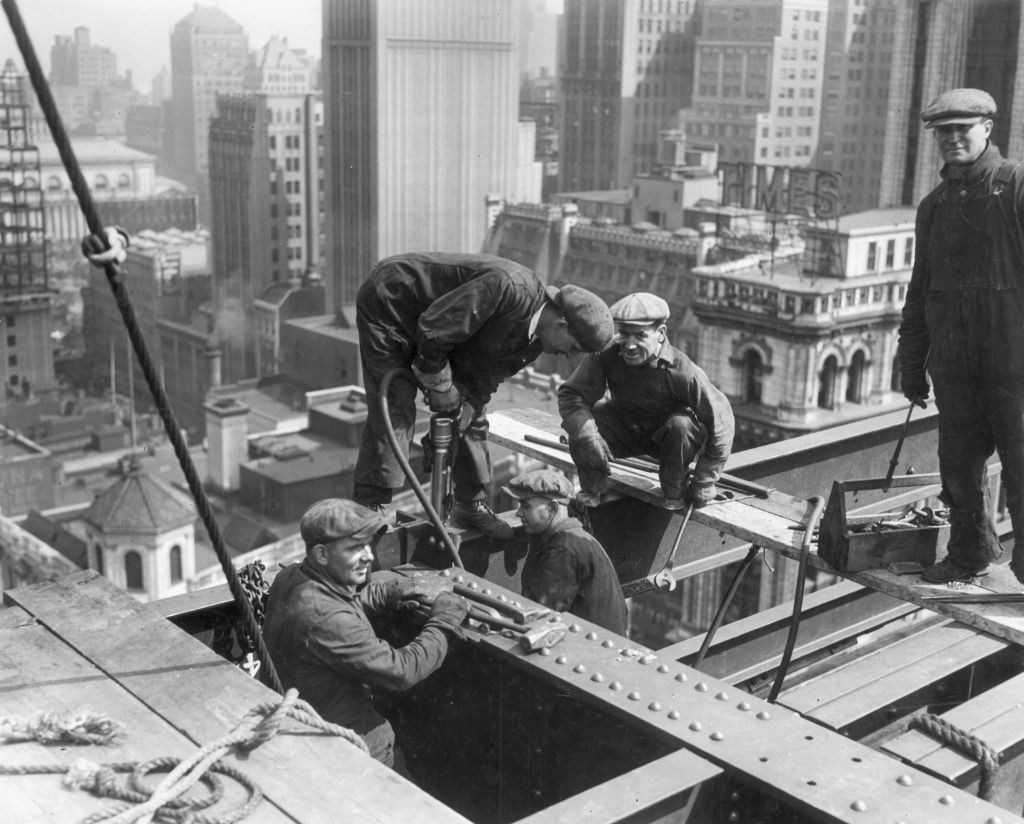 Five construction workers in 1930s attire building a high-rise structure in a densely built-up urban area, showcasing the architecture and skyline of the time period