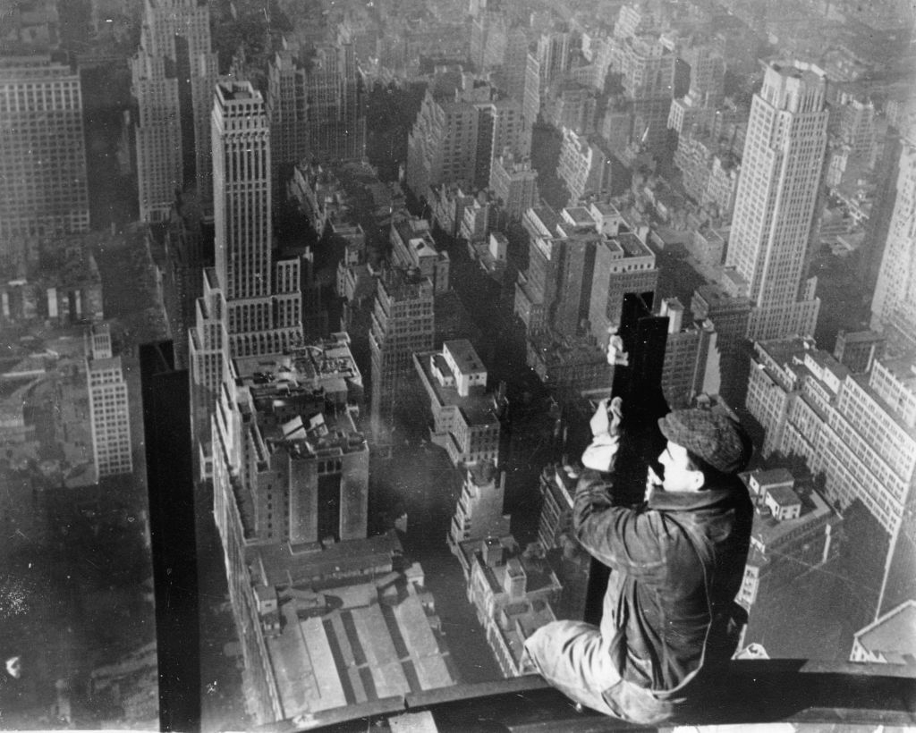 A construction worker sits on a steel beam high above New York City, with skyscrapers and buildings visible below