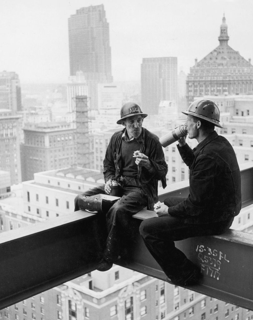 Two construction workers sit on a steel beam high above a city, eating lunch and drinking, with skyscrapers in the background