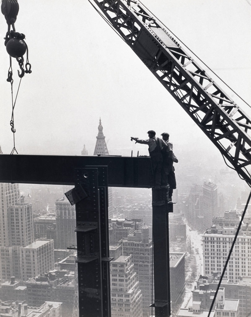 Two construction workers balance on a steel beam, high above the city skyline, with one pointing into the distance, while a large crane looms above them