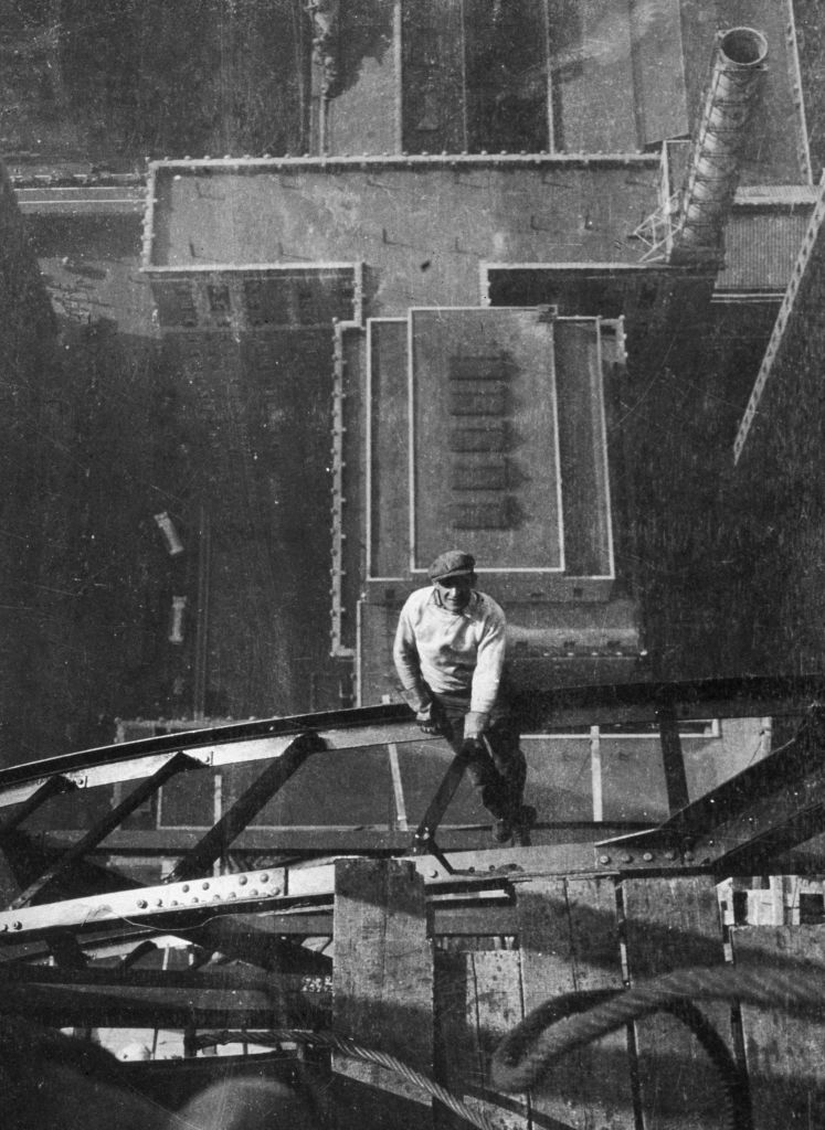 A construction worker stands on a high steel beam of a skyscraper under construction, with buildings and a cityscape visible far below him