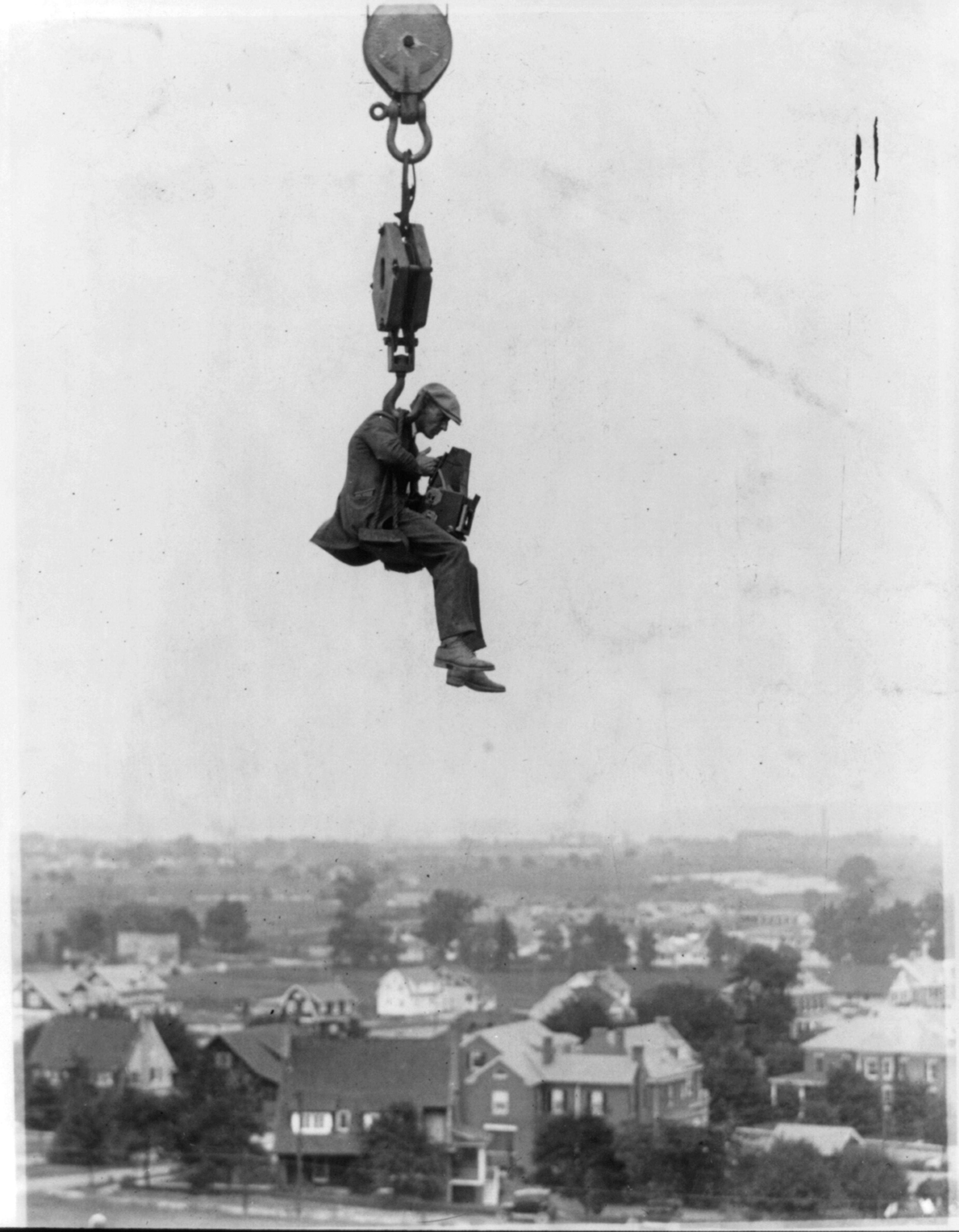A man is suspended high in the air by a crane over a town, holding onto a camera, capturing the scene below