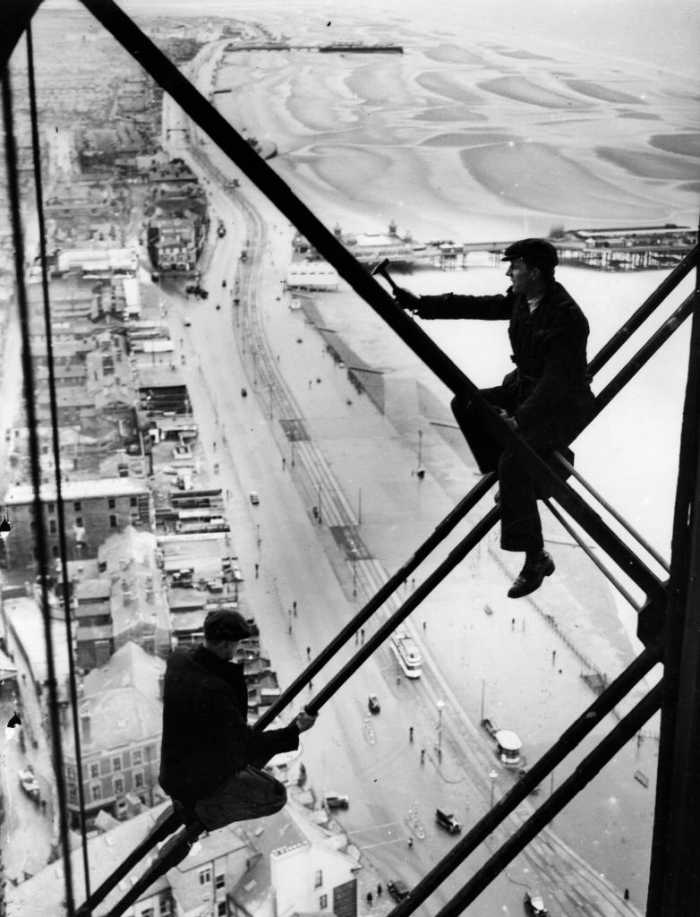 Workers perched on metal beams high above a city street, engaging in construction work on a building framework overlooking a beach