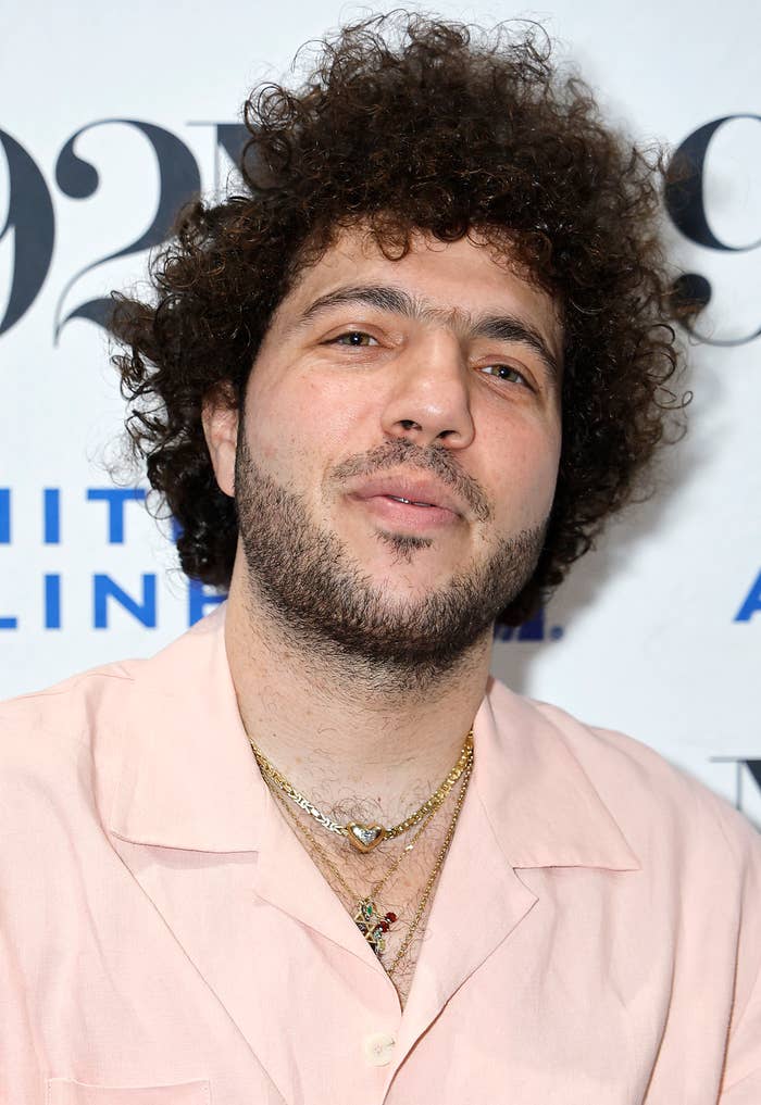 Benny Blanco at an event, wearing a pink shirt and multiple gold necklaces, standing in front of a backdrop with text