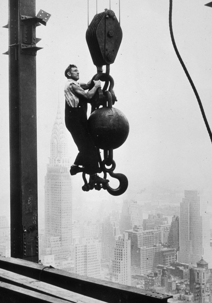 Man hanging from construction crane hook above city skyline with skyscrapers in background