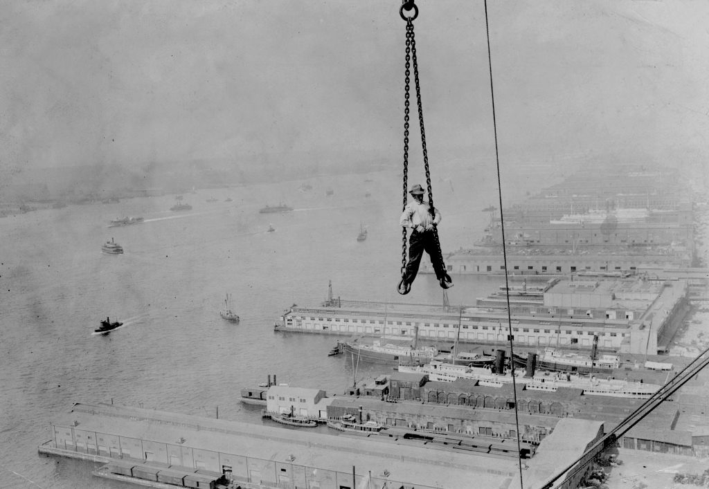 A man hangs from a crane over a dockyard, elevated high above the ground with ships and buildings visible in the background