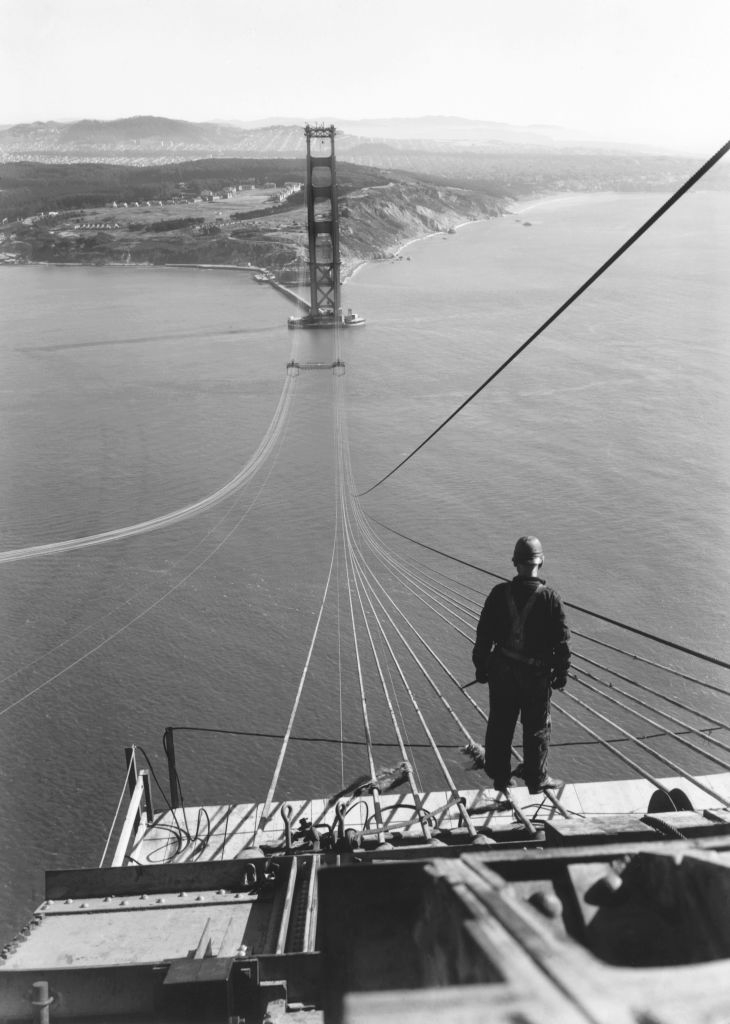 A construction worker in the foreground looks towards the incomplete Golden Gate Bridge in the distance. Cables extend from the foreground towards the bridge