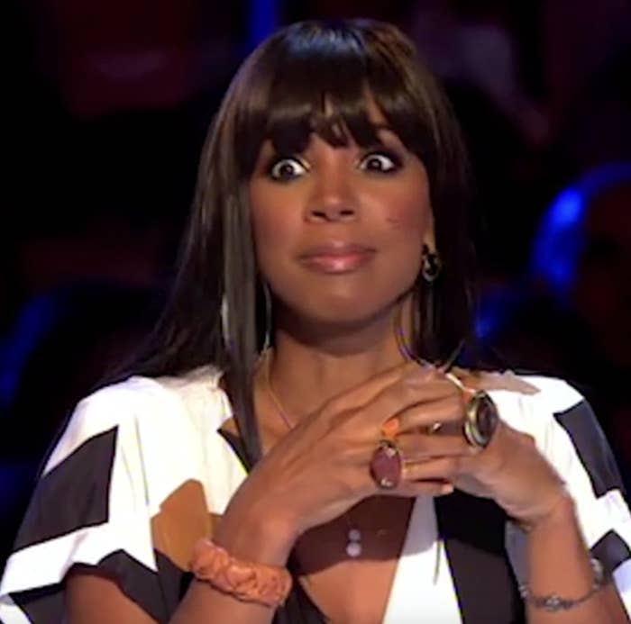 Kelly Rowland sits at a judging table, hands clasped with wide eyes, wearing a geometric-patterned blouse and statement rings