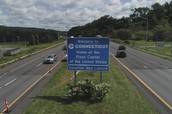 “Welcome to Connecticut” highway sign, reading “Home of the Pizza Capital of the United States. Governor Ned Lamont” with cars on the road in the background