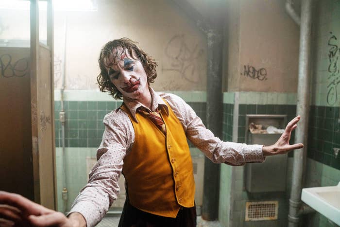 Joaquin Phoenix dressed as the Joker, wearing a mustard vest, white shirt with patterns, and red tie, performs an expressive dance in a grimy bathroom