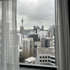 solo trip to japan itinerary View of Auckland city skyline through a window with sheer curtains, showing tall buildings and the Sky Tower in the background