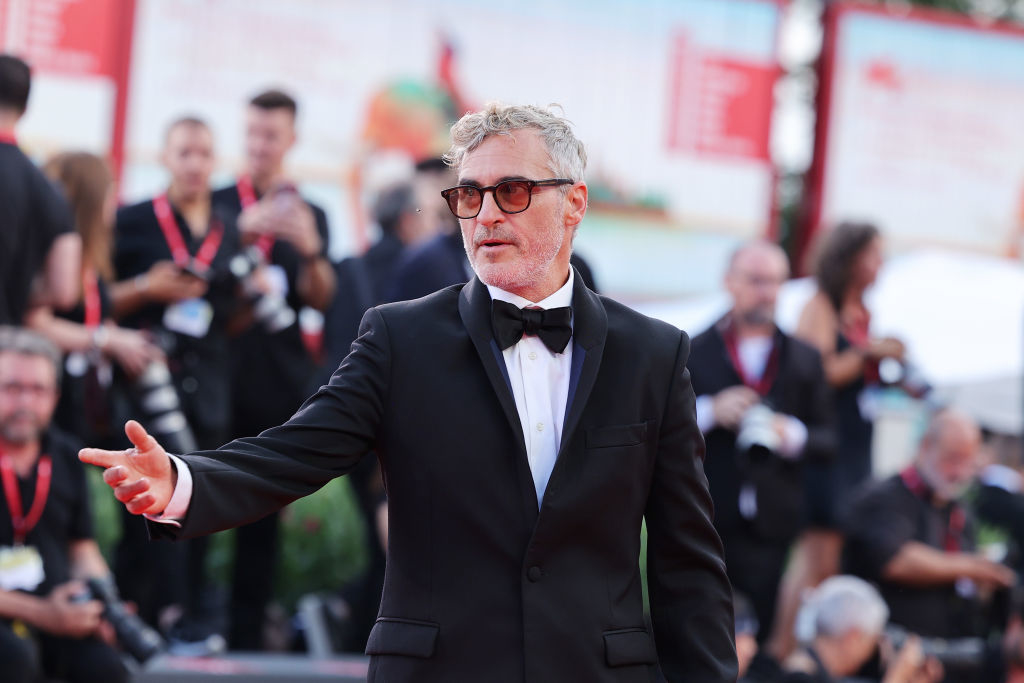 Joaquin Phoenix on the red carpet, wearing a black tuxedo and sunglasses, extending his hand. Background shows a busy, out-of-focus event setting