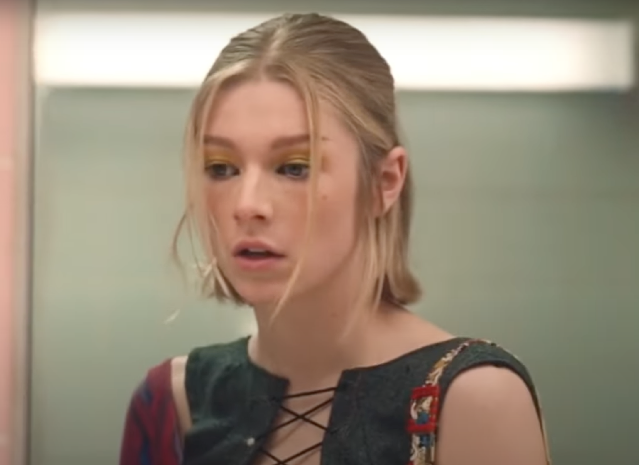 Hunter Schafer with a neutral expression, dressed in a stylish outfit featuring crisscross lacing at the front, looks off-camera indoors