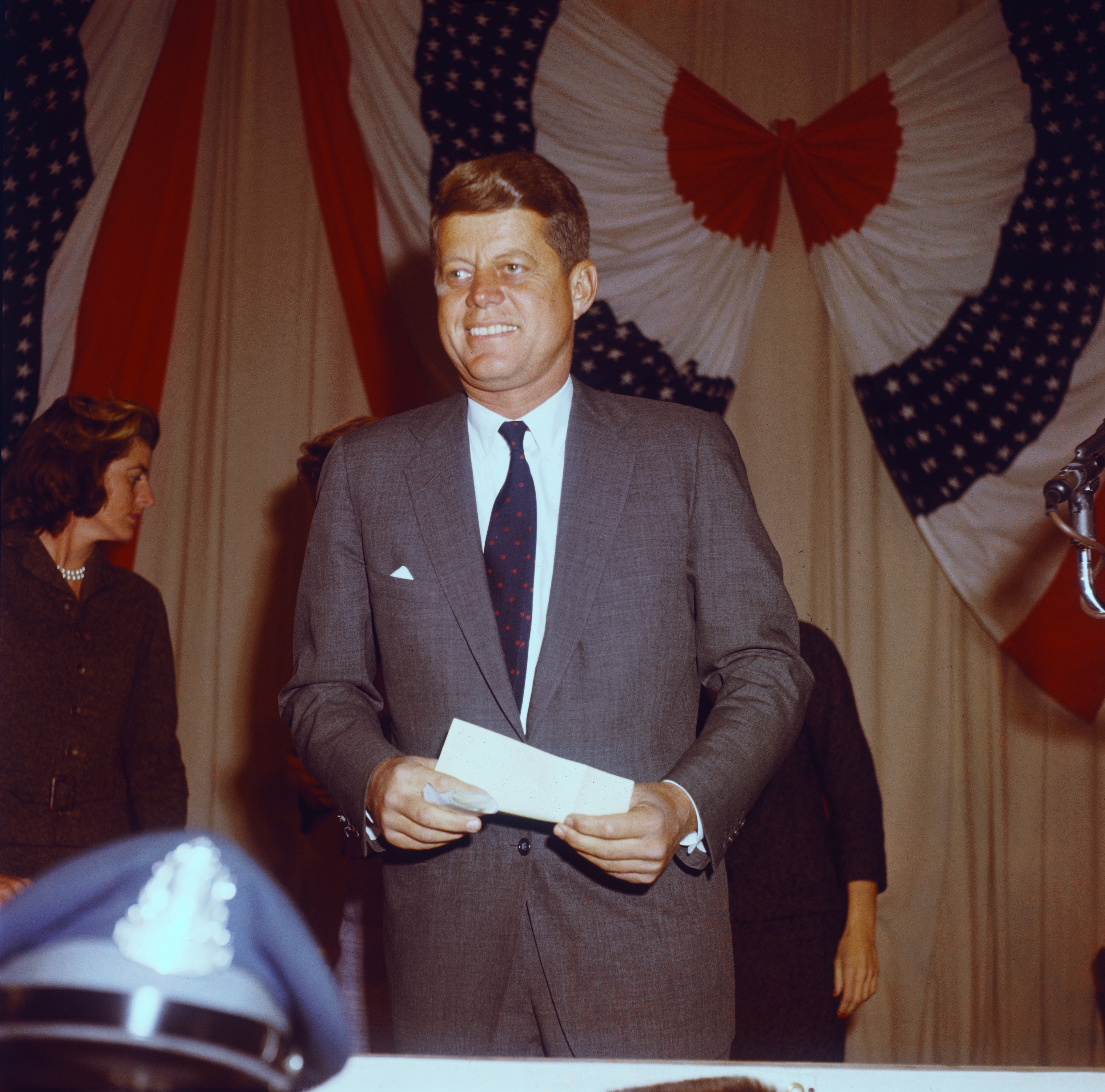 John F. Kennedy, in a suit and tie, stands smiling and holding a piece of paper. Draped American flags are visible in the background. A woman and a police hat are in the foreground