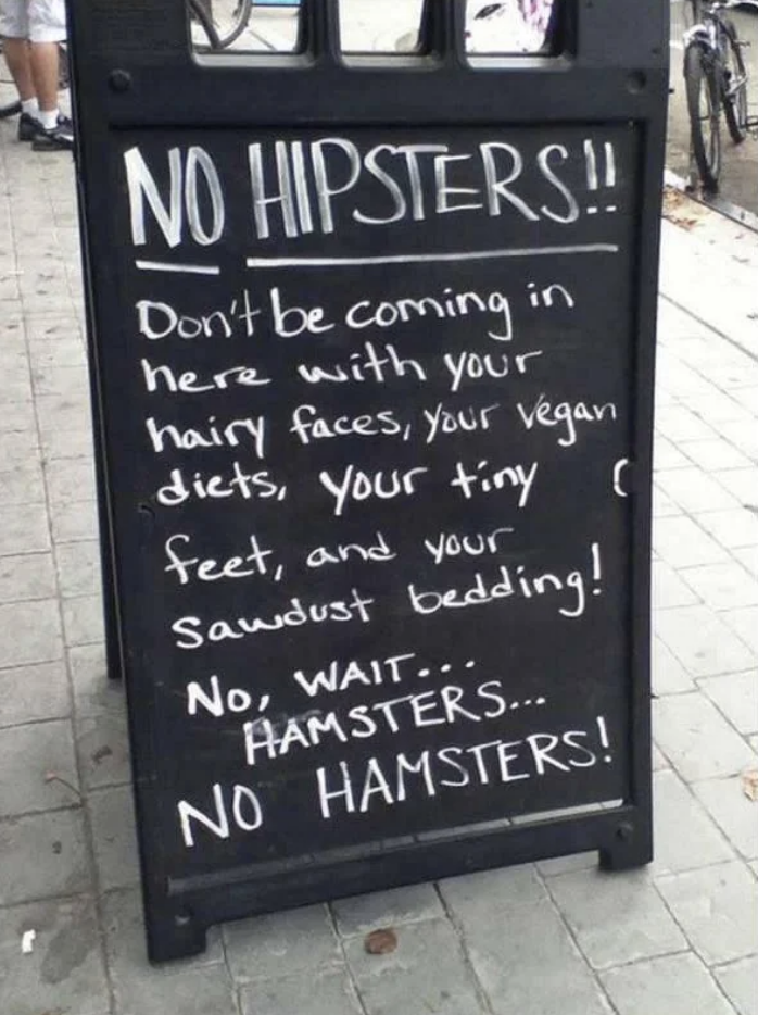A sidewalk sign reads: "NO HIPSTERS!! Don't be coming in here with your hairy faces, your vegan diets, your tiny feet, and your sawdust bedding! No, wait… HAMSTERS… NO HAMSTERS!"