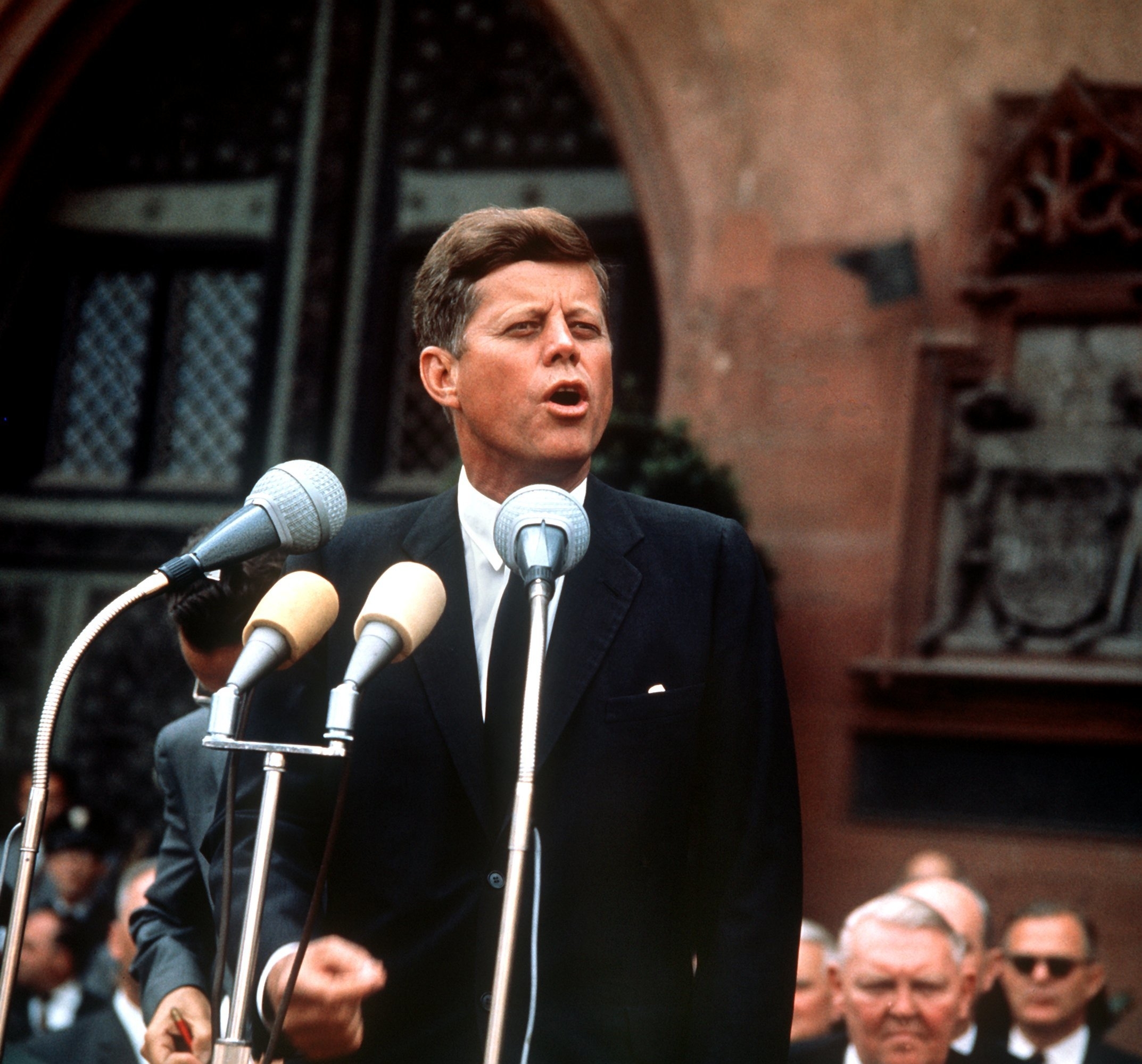 John F. Kennedy speaks passionately at a microphone, dressed in a suit and tie. A crowd is visible behind him