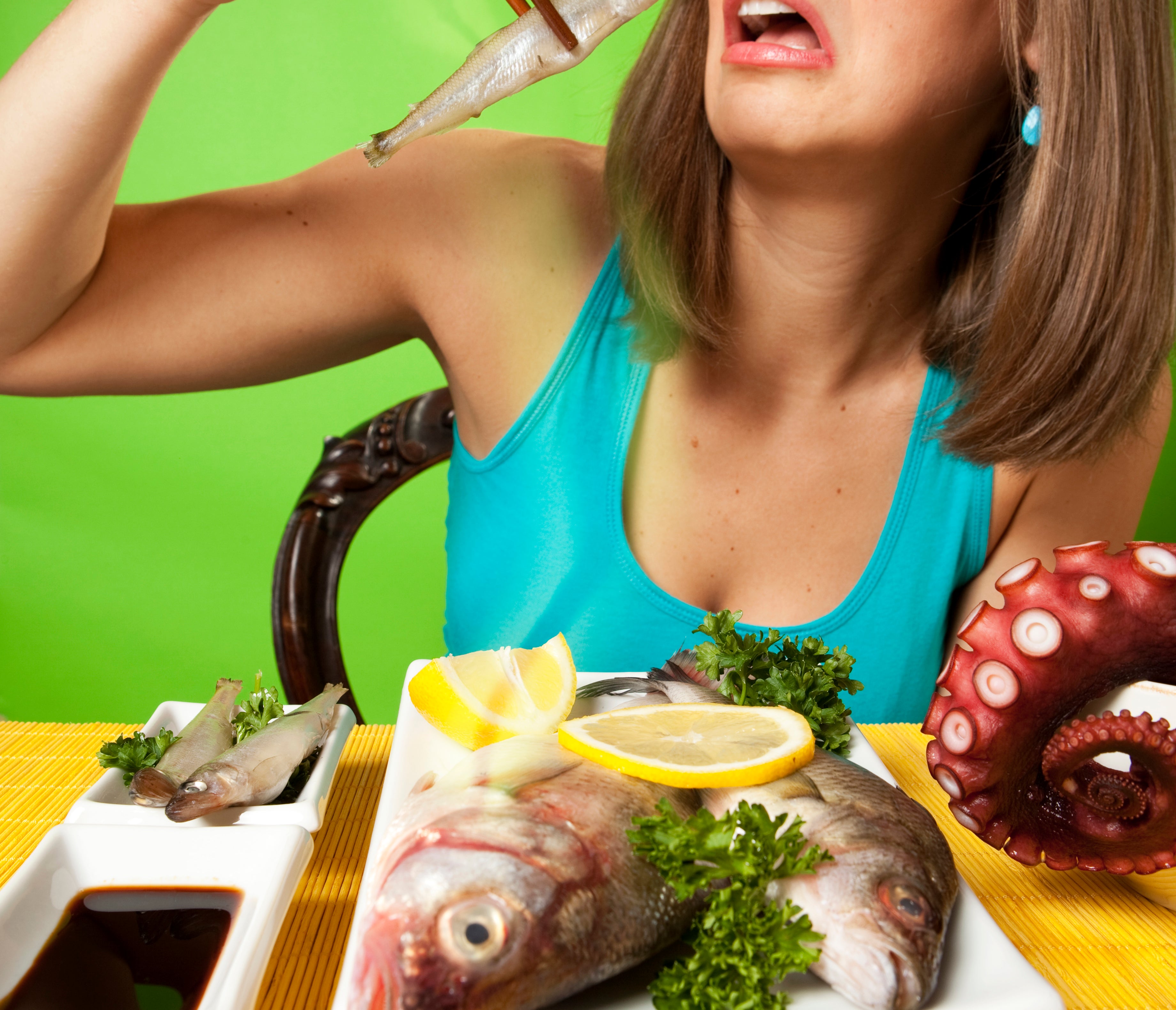 A woman in a blue top holds a small fish with chopsticks, looking uneasy, with a plate of seafood (fish, lemon, parsley, octopus) in front of her
