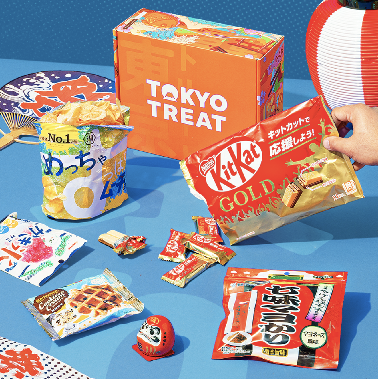 An assortment of Japanese snacks from TokyoTreat, including chips, KitKat Gold, Pocky, and various candies, displayed on a blue surface with a Japanese lantern and fan