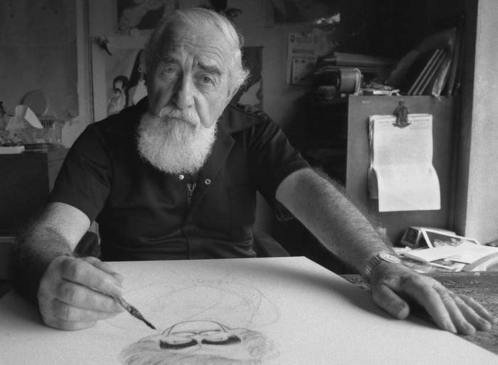 An elderly man with a white beard, dressed in a short-sleeved shirt, is sketching on a large piece of paper. He appears focused on his work