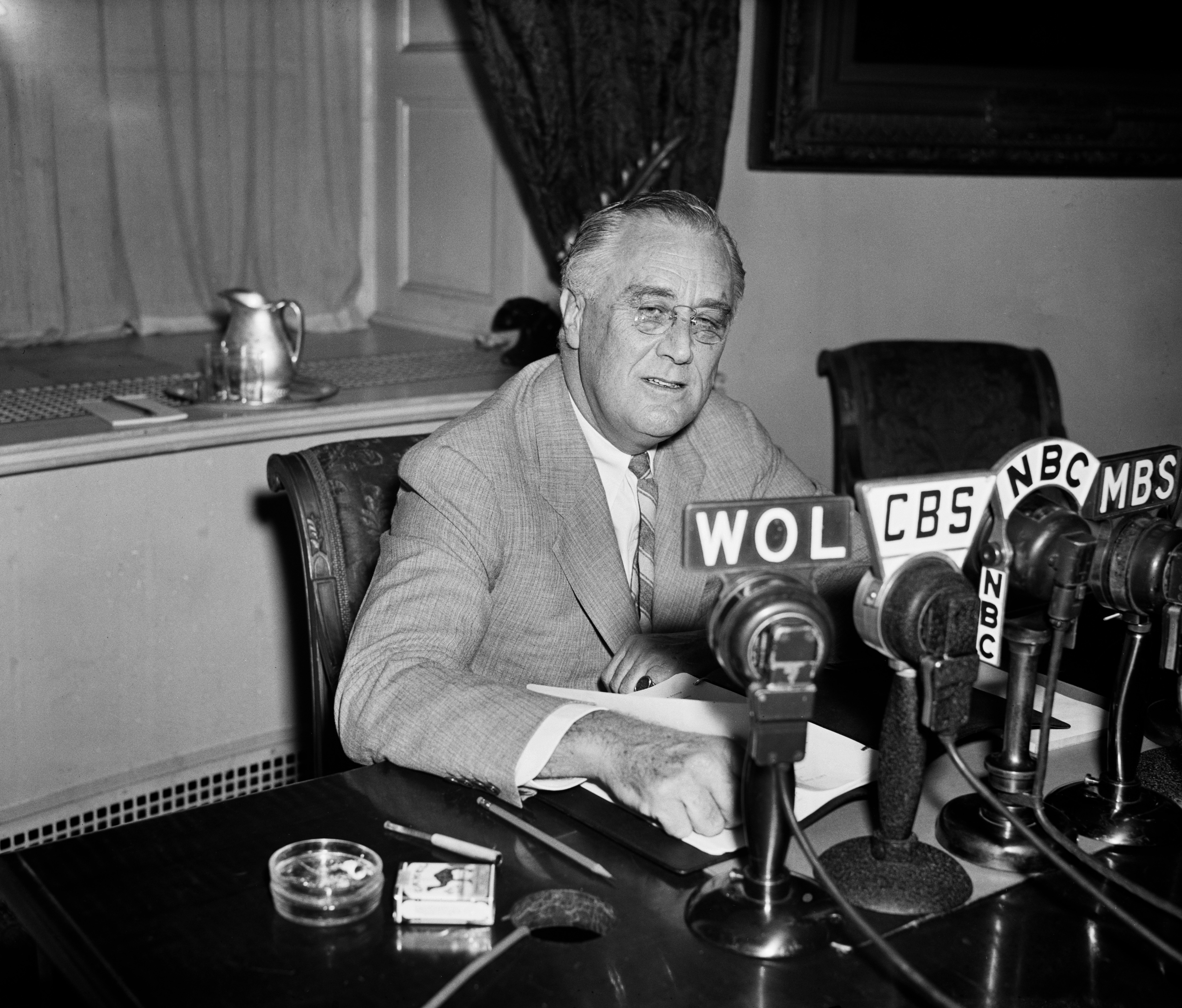 Franklin D. Roosevelt sits at a desk with several microphones in front of him, including WOL, CBS, NBC, and MBS