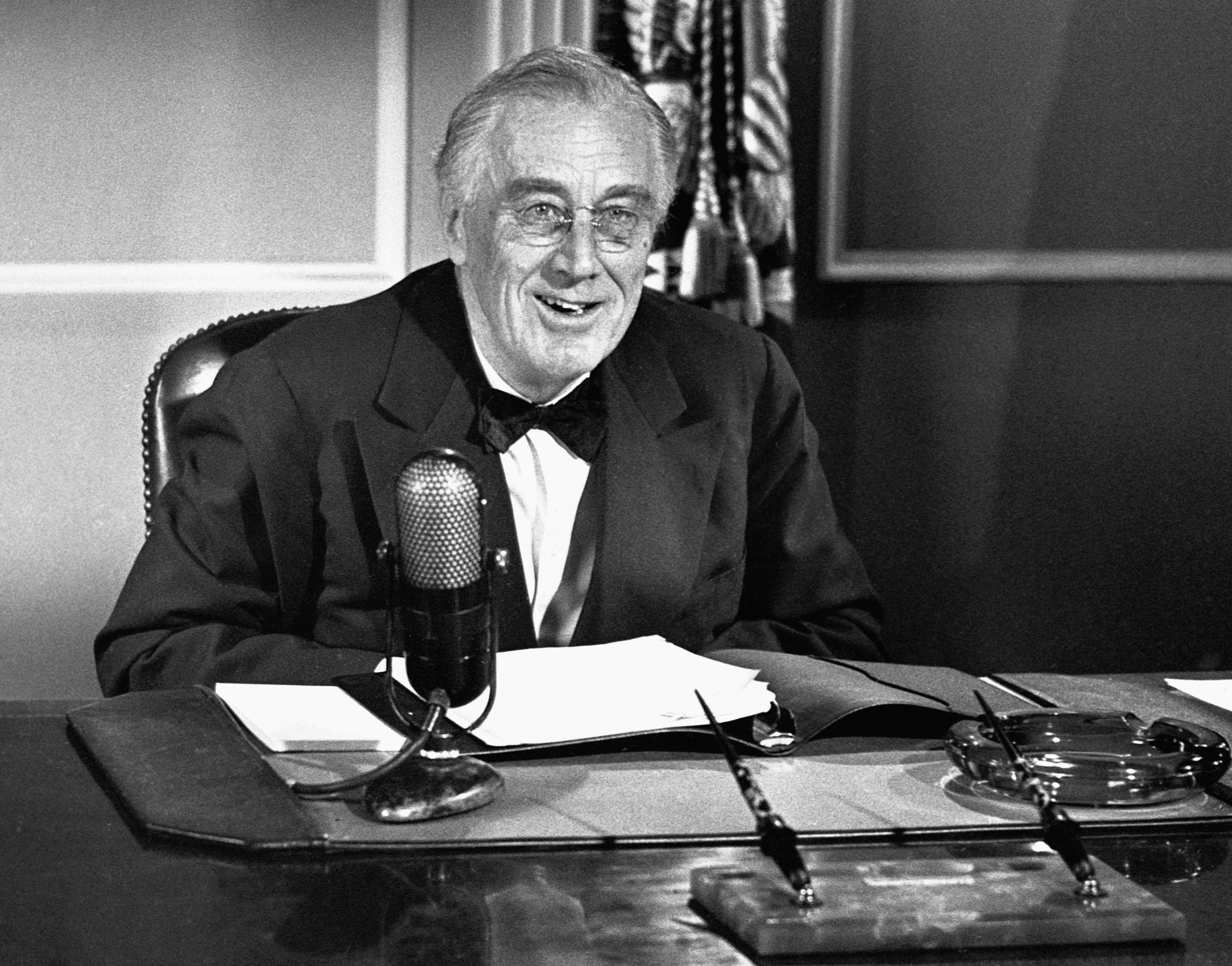 Franklin D. Roosevelt sits at a desk with a microphone, delivering a speech, smiling. Papers and pens are on the desk in front of him