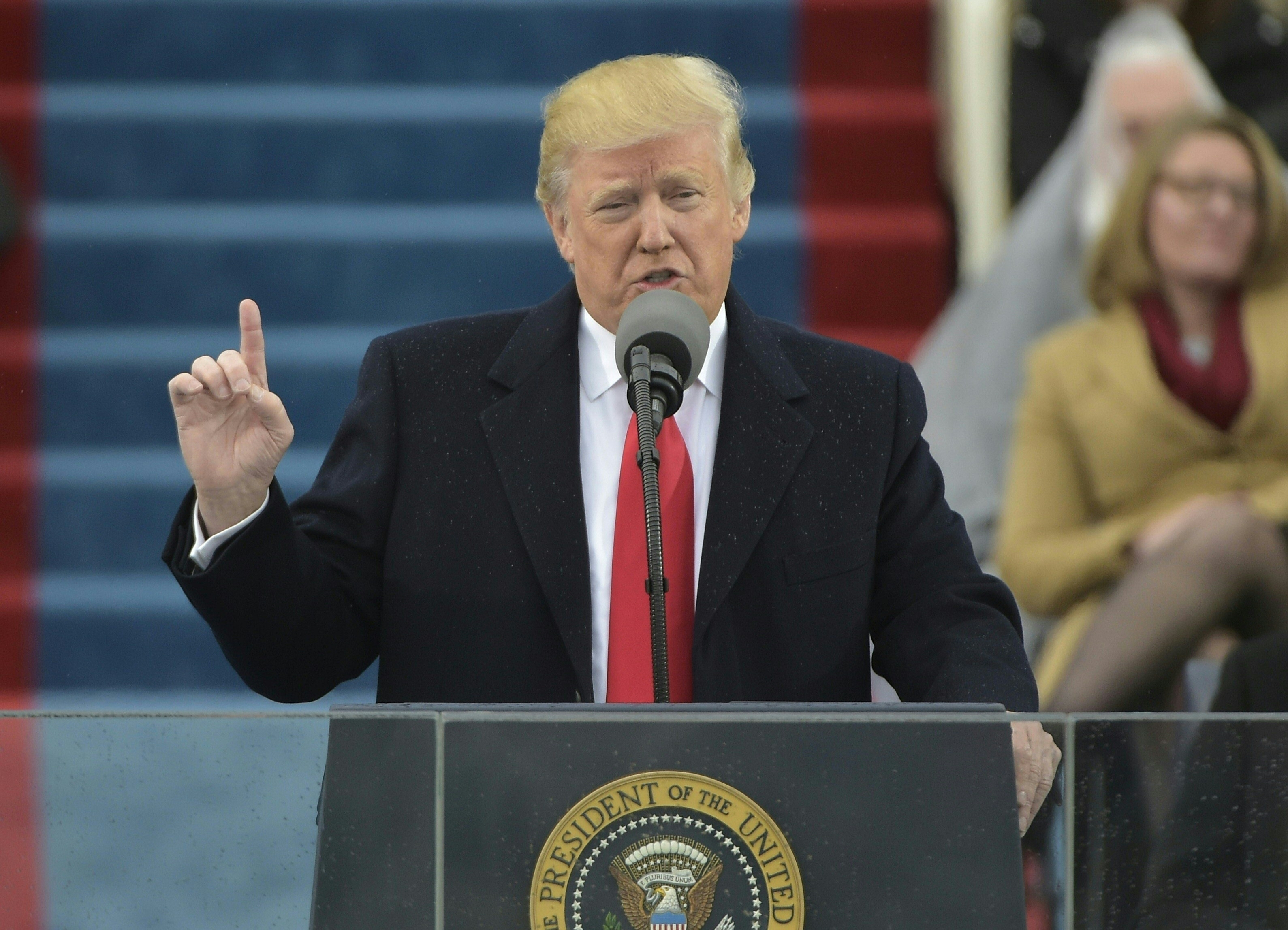 Donald Trump speaks at a podium during an outdoor event. He is wearing a suit and overcoat, and is gesturing with his right hand