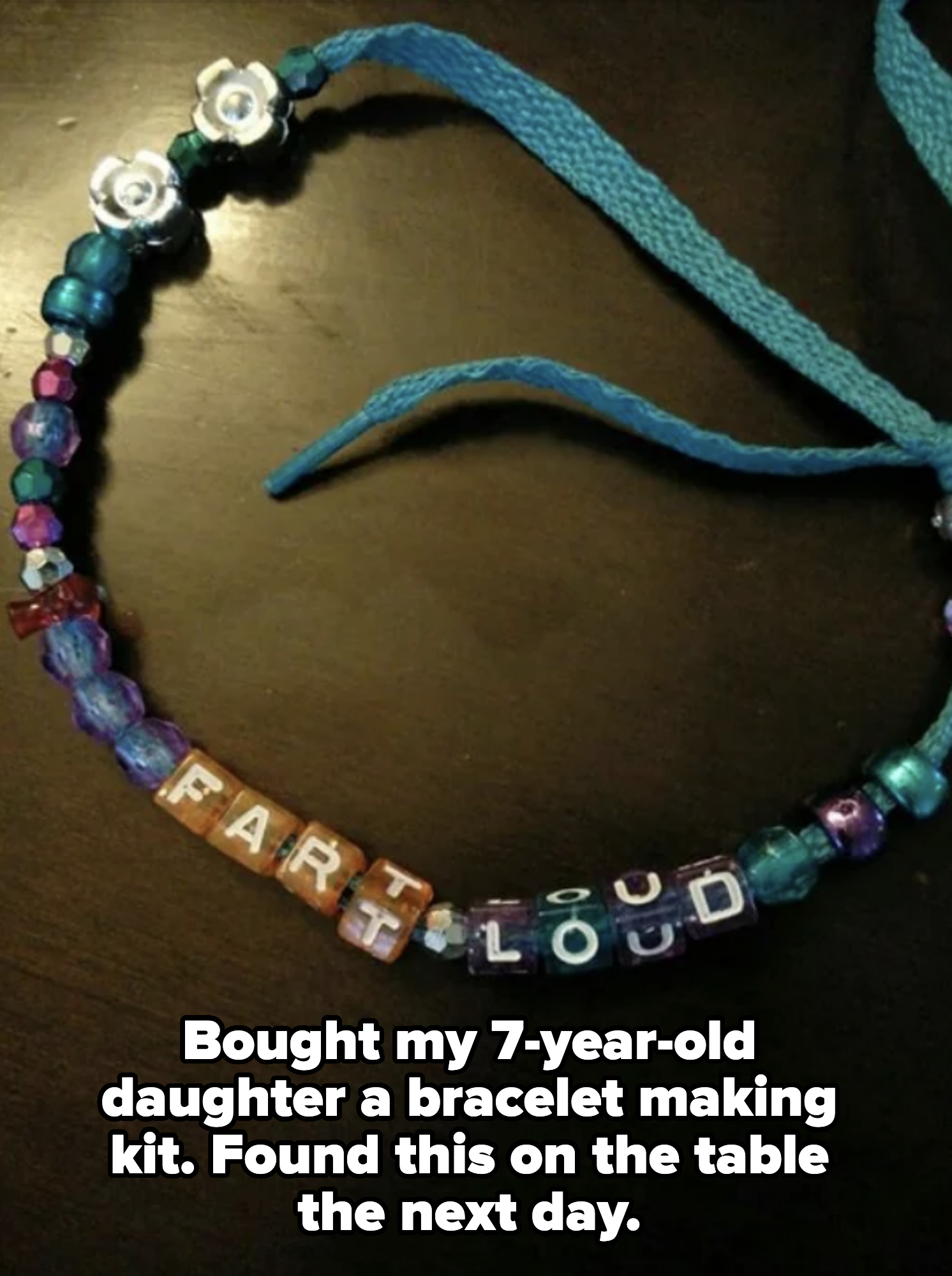 A handmade beaded necklace spells out "FART LOUD" with letter beads, among other blue and purple beads, tied with a blue ribbon