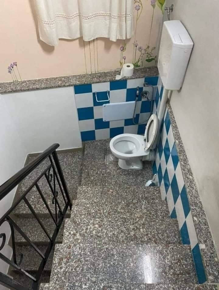 A restroom with a toilet and tiled wall located at the bottom of a stairway. There is a roll of toilet paper on the ledge above the toilet