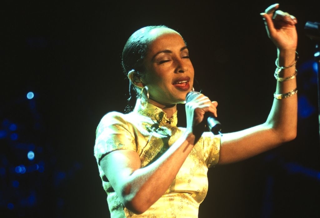 Sade performing on stage, wearing a stylish dress with her hair slicked back, holding a microphone and singing with eyes closed