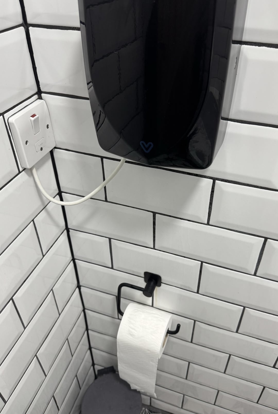 Public restroom wall with white tiles, a black hand dryer, and a toilet paper holder with a roll of toilet paper