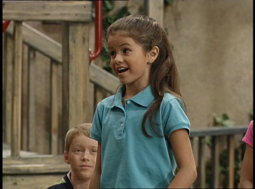 Selena Gomez standing and speaking while a young boy with light hair and seated in the background listens attentively