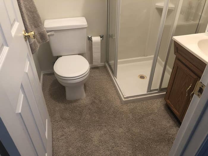 A small bathroom with a glass shower, white toilet, white sink on a wooden cabinet, and carpeted floor