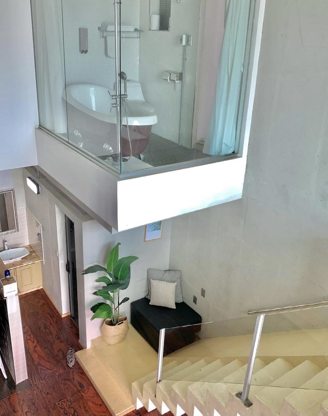 Modern, multi-level interior with a glass-walled bathroom overhanging above a sitting area with plant and chair, and a staircase leading downstairs