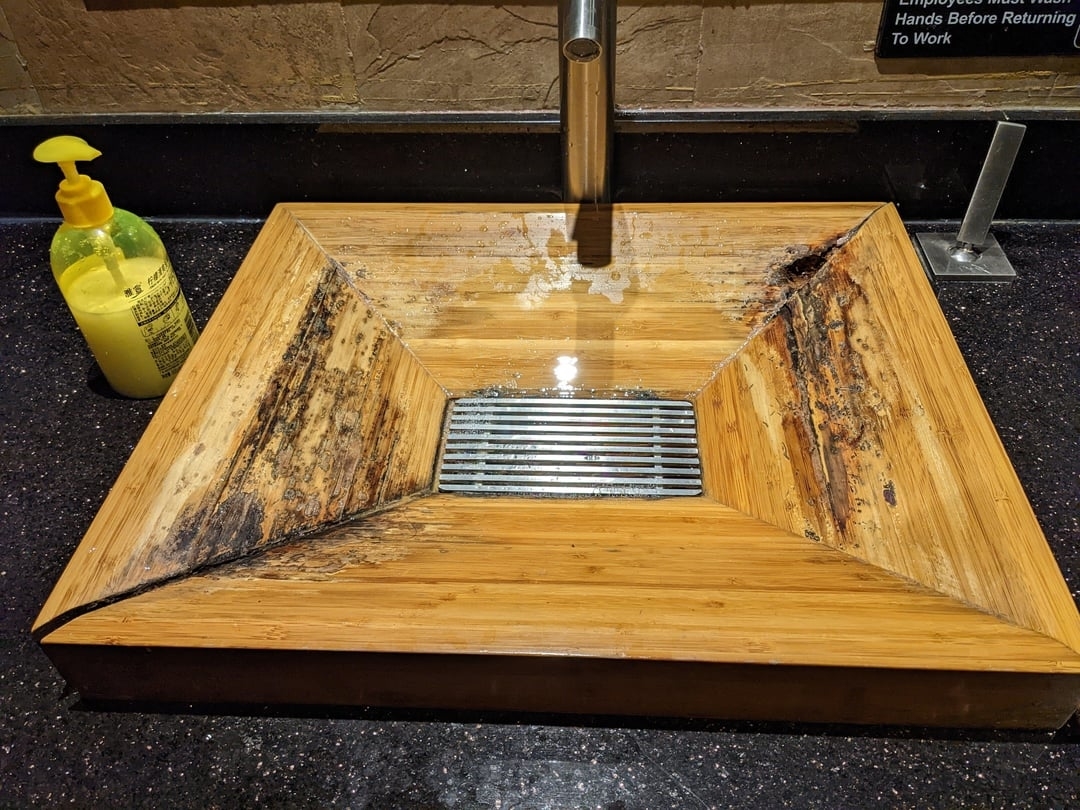 Wooden sink with a sloping design showing visible mold and water damage, with a yellow liquid soap dispenser on the left