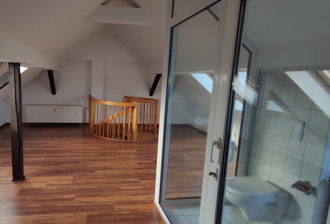 A spacious attic room with a wooden floor, a spiral staircase, and a small bathroom with a toilet behind glass doors