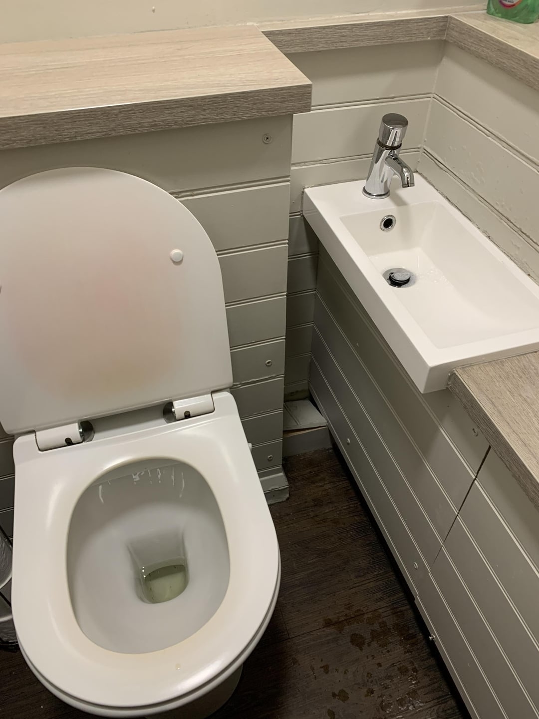 A small bathroom setup with a toilet and a sink placed close together, both fitted into a corner with wooden paneling on the walls