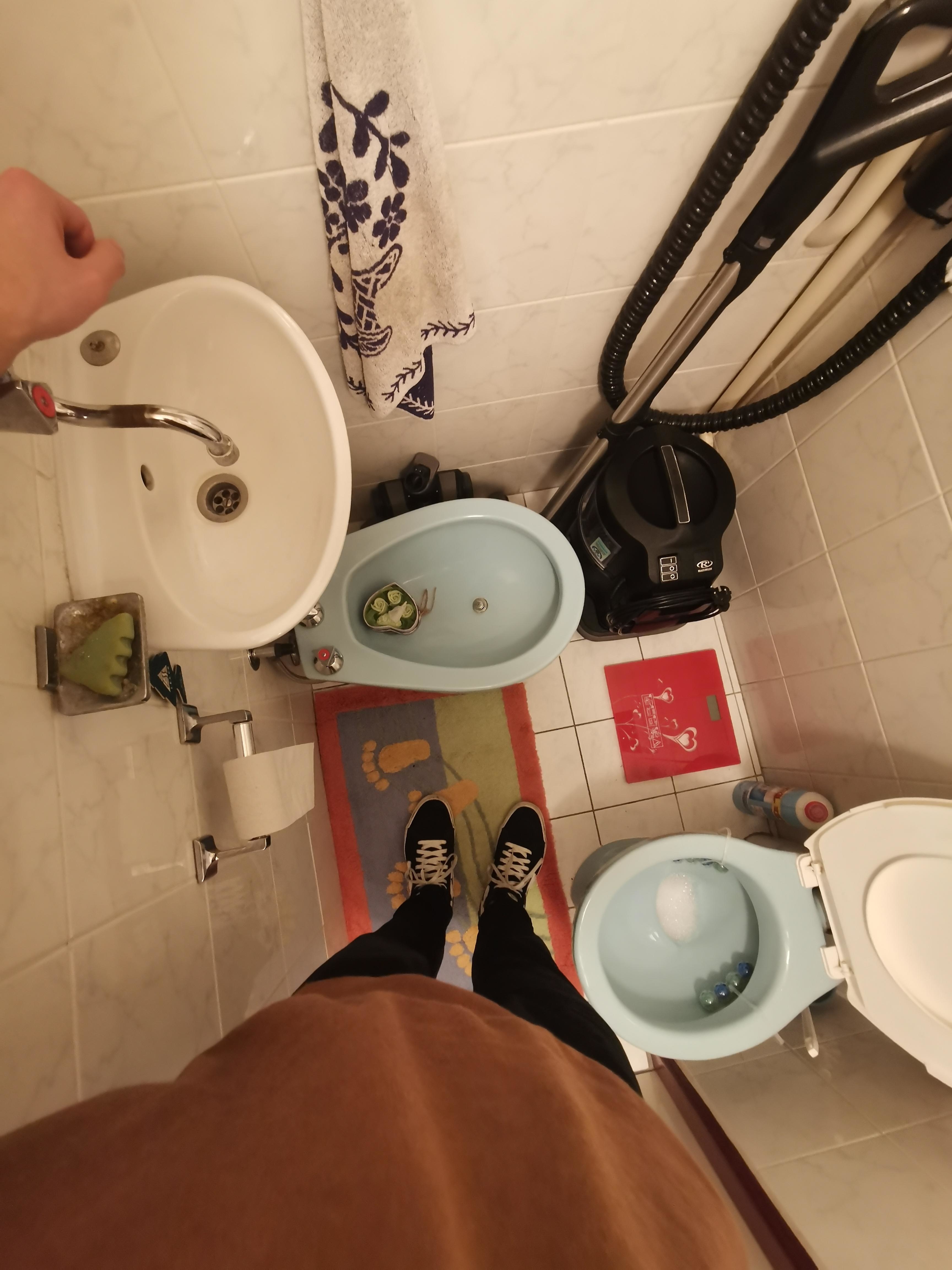 Person standing in a bathroom near a sink, toilet, bidet, and vacuum cleaner on the right side