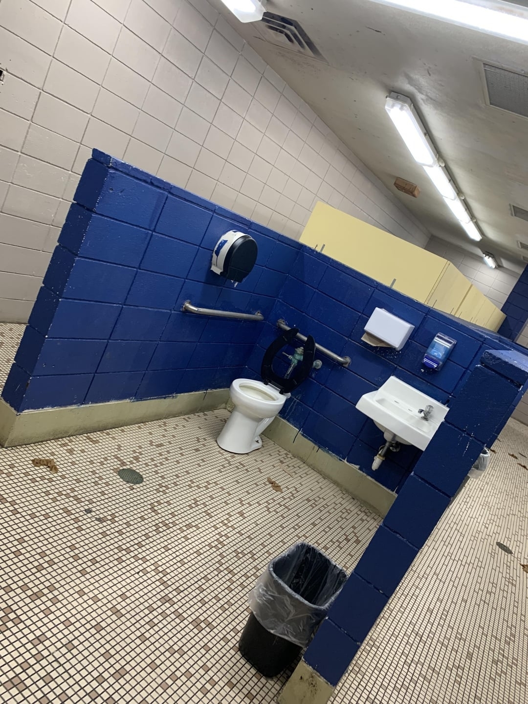 Public restroom with a toilet, sink, soap dispenser, paper towel dispenser, and trash can. The walls are made of tiles and the floor has small square tiles