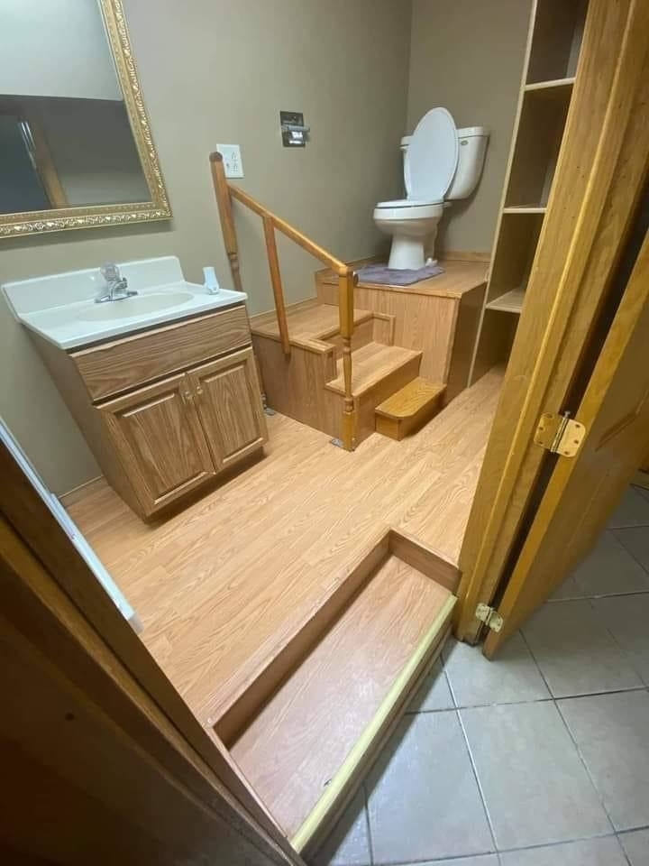 A bathroom with a sink and vanity next to a toilet elevated by stairs and railings. The design seems unconventional, raising accessibility concerns