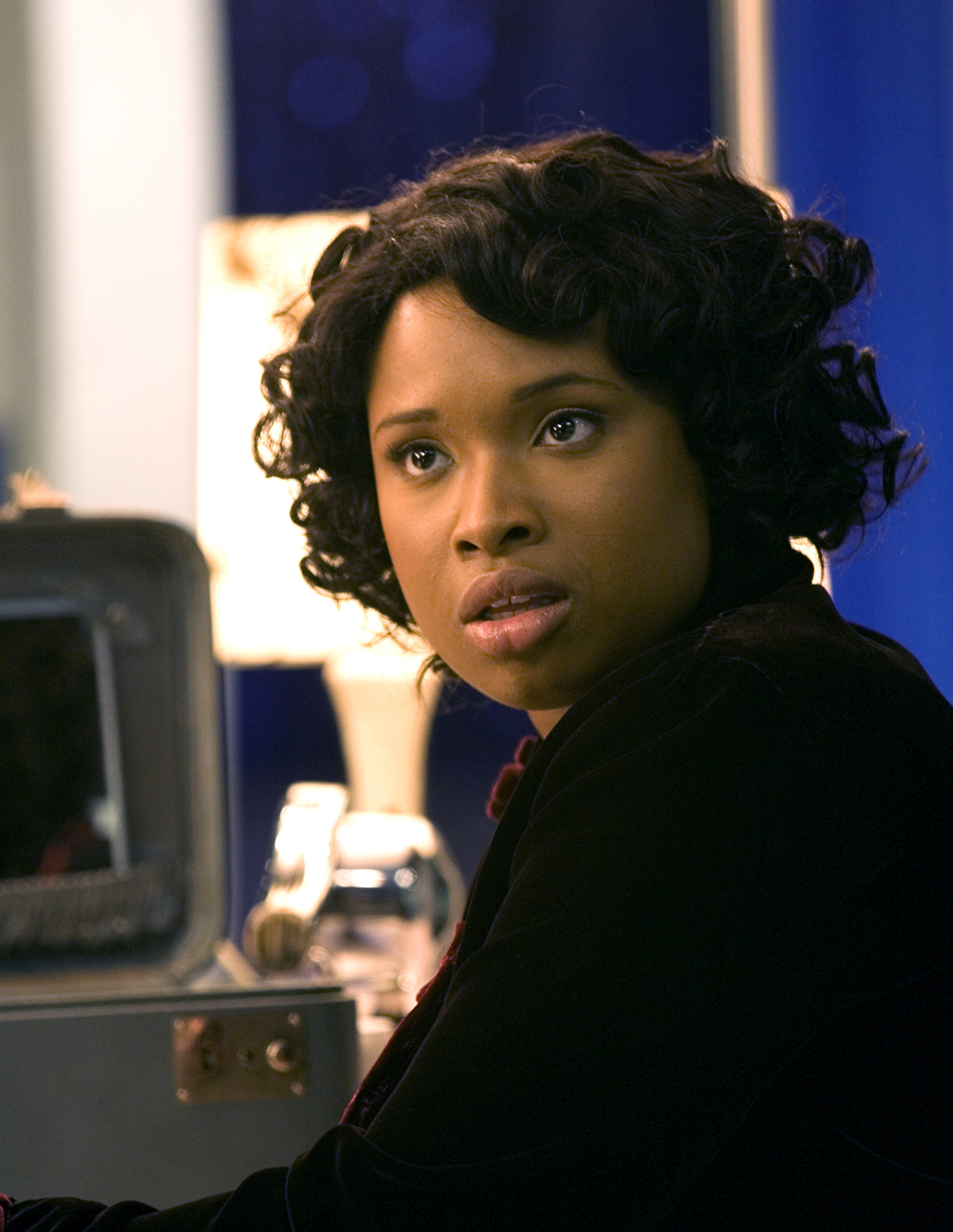 Jennifer Hudson, with short curly hair, is seated in a room looking to the side in a thoughtful expression, with a vanity mirror behind her